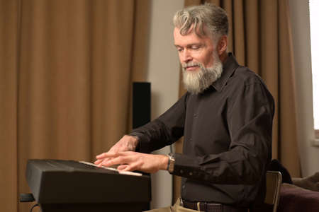 adult solid gray-haired man with a stylish beard plays the electronic piano at home in his spare timeの写真素材