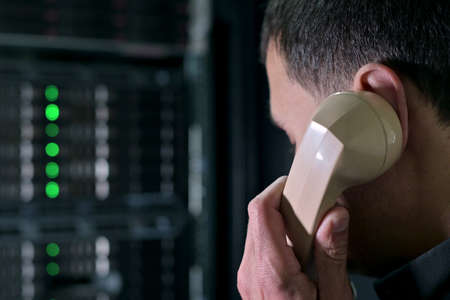 A man calls on an old phone against the backdrop of a modern server cabinet. A comic image of the mixing of eras of technology. Obsolescence of technologyの写真素材