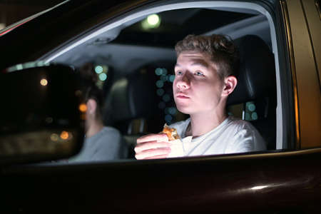 Young guy eating burger in the car driving late at night in the parking lot. Close up, view through windowの写真素材
