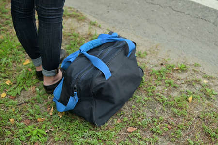 a woman is waiting for a ride on the side of the road, a travel bag lies nearby, close upの写真素材