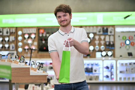 Young salesman in electronics store puts goods into paper bag and holds out