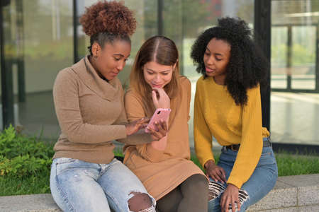 Three black and white female friends chatting in the park looking at photosの写真素材