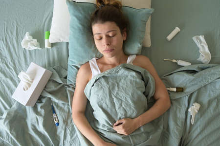 A sick-looking young woman lies and suffers in bed. Medicines are scattered aroundの写真素材