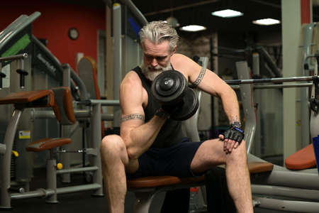 A gray-haired man with a beard and tattoos pumps biceps in the gym