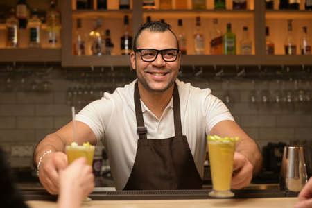 smiling black waiter serves cocktails to customers at the barの写真素材