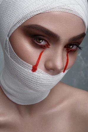 Horror female Patient with Blood Drops from Eyes and white Bandage on Head and Mouth. Halloween Makeupの写真素材