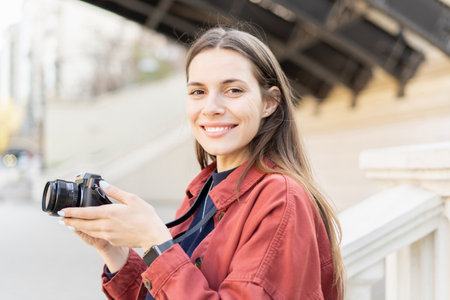 Side close up of a cheerful young photographer girl, looking in the camera smiling and holding her...