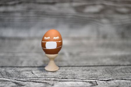 A brown chicken egg in mask in holder on the wooden background. Coronavirus Covid-19. Concept photo....