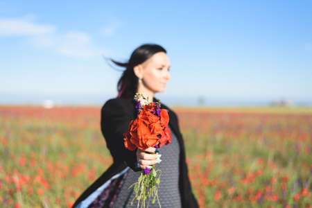 A concept portrait of defocussed beautiful young darkhaired woman with bouquet in focus in the foreground on the poppy field.の写真素材