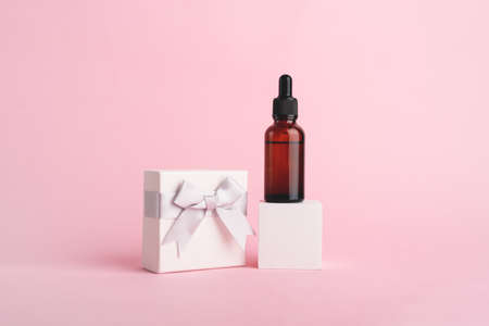 Serum glass bottle with pipette on the white cube and white gift box with silver ribbon bow near on pink background. Natural Organic Spa Cosmetics. Trendy concept. Front view.の写真素材