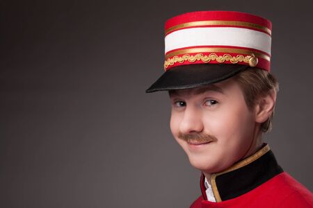 Portrait of a concierge (porter) in a red jacket on a grey background.