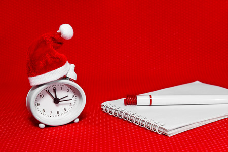 The clock in Christmas hat, notebook and pen on a bright red background. Christmas still-life...