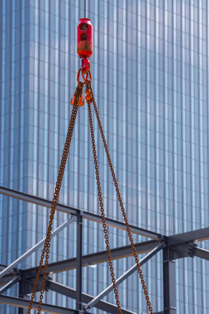 Crane hoisting block with hook on steel chain on steel rope. Glass facade of a modern skyscraper in the background. Loading\unloading of building materials on construction building site.の写真素材