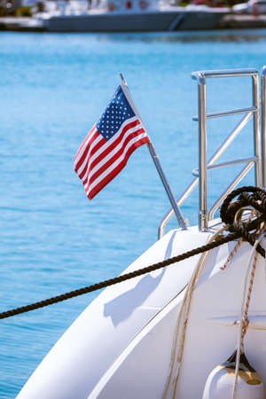 The flag of the United States flutters in the wind on a stainless steel flagpole at the stern of a motor yacht. Marina in the port city in beautiful summer sunny weather.の写真素材