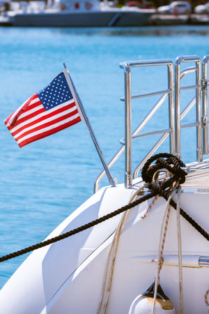 The flag of the United States flutters in the wind on a stainless steel flagpole at the stern of a motor yacht. Marina in the port city in beautiful summer sunny weather.の写真素材