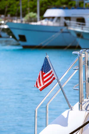 The flag of the United States flutters in the wind on a stainless steel flagpole at the stern of a motor yacht. Marina in the port city in beautiful summer sunny weather.の写真素材