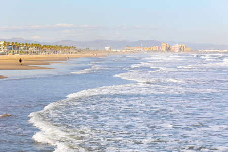 Summer vibes on the sunny autumn beach of Malvarrosa in Valencia, Spain. Vast expanses of smooth fine sand on the sea coast attract vacationers to solitary walks along the bubbling foamy waves.の写真素材