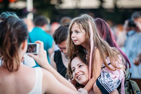 Yessentuki, Stavropol Territory / Russia - August 12, 2017: beautiful pretty girl child blonde with pink hair sitting on the shoulders of  mother, they are photographed and watching the concert outdoorsのeditorial素材