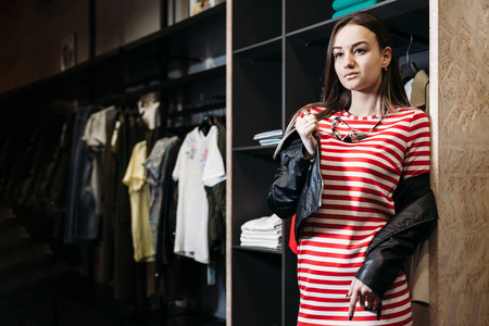 The young pretty woman choosing, trying and buys dresses at shop clothing. Banner for online store clothing. Girl posing in fashionable clothesの写真素材