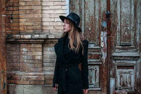 Blonde beautiful girl with long hair, in black coat in hat, stands on the background of vintage antique old wooden door old brick house. For concept design.の写真素材