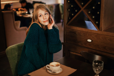 Beautiful young woman blonde drinks coffee in a restaurant. surprised emotion with the phone. coquetteの写真素材