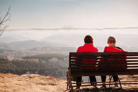 couple sitting on bench at sunset, looking at beautiful view from mountain to mountainの写真素材