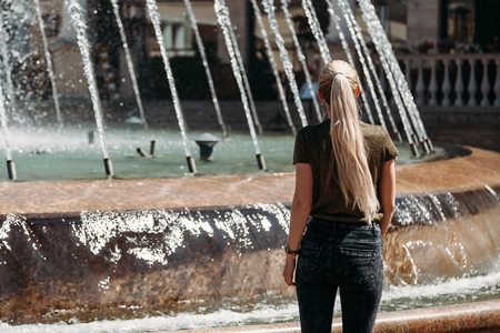 Young girl with long hair in jeans near the fountain, in summer, view from the backの写真素材