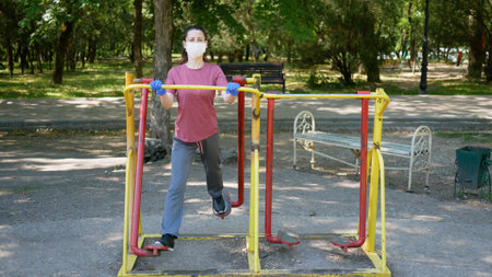 Young woman in gloves and face mask sports on outdoor exercise equipment in Park. Protection from...