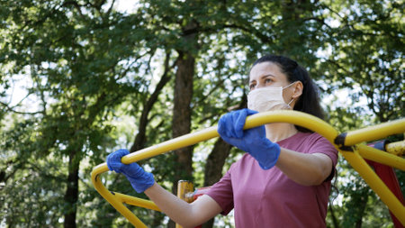 Young woman in protective face mask and gloves, goes in for sports in outdoor exercise equipment in...