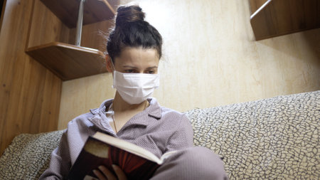 Young woman with protective face mask reading a book in home self isolation quarantine. Coronavirus pandemic COVID-19. Stay home.の写真素材