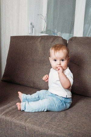adorable baby boy sitting on couch with fingers in his mouthの写真素材