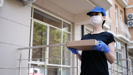 Courier, delivery man in protective mask and medical gloves delivers small box parcel. Delivery service under quarantine, disease outbreak, coronavirus covid-19 pandemic conditions. Stay home.の写真素材