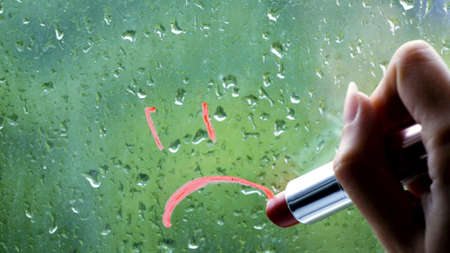 concept of bad weather on street. young woman draws sad smiley on window, outside window rain.