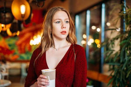 Young blonde woman holding a paper cup of hot coffee of takeaway in her hands indoors in cafe and...