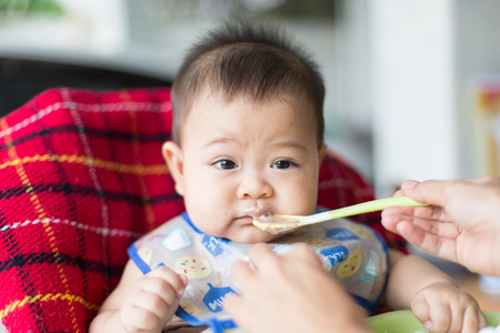 the baby learns to eat by himself. he can use spoon well. so he is very happy but his mother still feed him (focus at his face)の写真素材