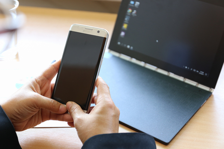 hands holding smart phone before laptop on wood desk backgroundの写真素材