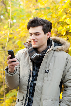 Young smiling man looking on his cellphone in autumn park.の写真素材