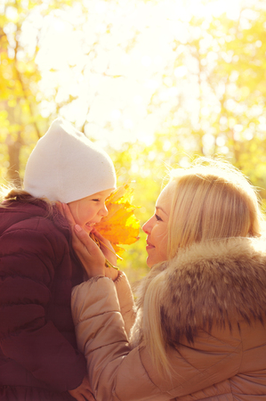 Happy mother holding her daughter's face in hands and smiling to her, happy childhood, mother's love, sunny backlight in autumn park.の写真素材