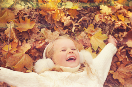 Little blonde girl portrait lying on autumn yellow maple leaves outstretched hands and smiling, play outdoor in autumn park, happy childhood.の写真素材