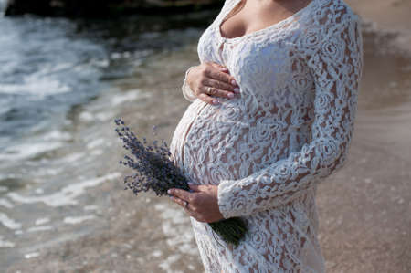 Future mother belly, pregnant woman posing near ocean in white lace dress, no faceの写真素材