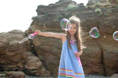 Little cute girl playing blowing soap bubbles, happy childhood concept, outdoor photo against seaの写真素材