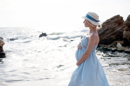 Happy future mother standing near sea, calm pregnant woman dressed in blue dress and hat