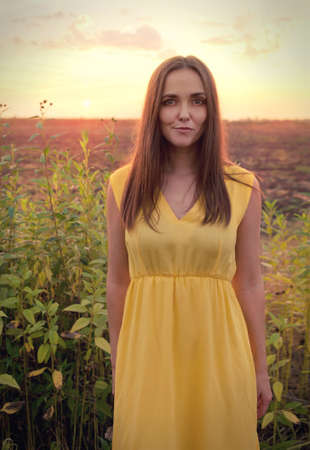 Beautiful young woman portrait against sunset evening field, woman dressed in yellow dress have a...