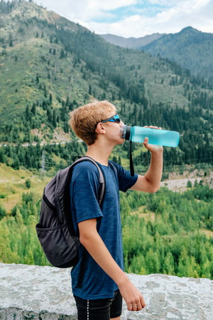 Young traveller stands on the backdrop of a stunning view with green mountains and drinks clear water from a blue bottleの写真素材