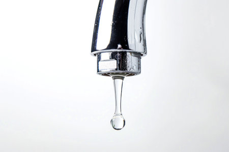 A close-up of a chrome tap with a single water droplet about to fall, set against a white background.の素材