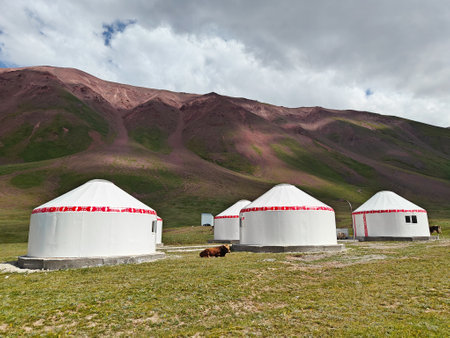 Traditional yurts are nestled in a green valley with mountains soaring in the background on a clear...