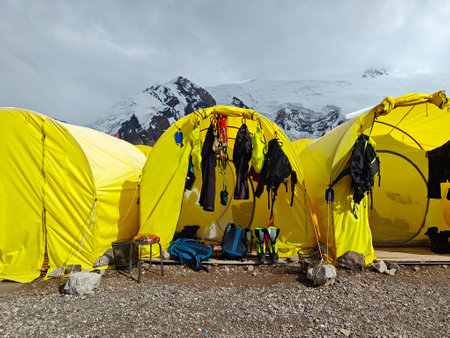 Bright yellow tents are pitched on rocky terrain with snowy peaks looming in a misty atmosphere as...