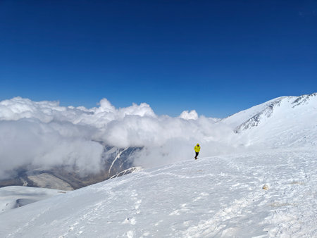 A lone mountaineer in a bright yellow jacket stands on a snowy slope of a peak, surrounded by clouds...