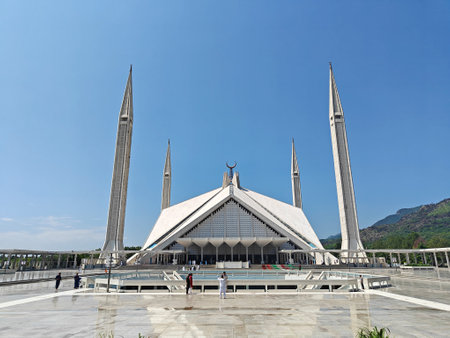 Shah Faisal Mosque in Islamabad with its iconic geometric design, tall minarets, and a vast courtyard under a clear blue sky, set against the backdrop of green Margalla Hills on a sunny day.の写真素材