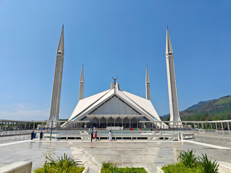 Shah Faisal Mosque in Islamabad with its iconic geometric design, tall minarets, and a vast courtyard under a clear blue sky, set against the backdrop of green Margalla Hills on a sunny day.の写真素材